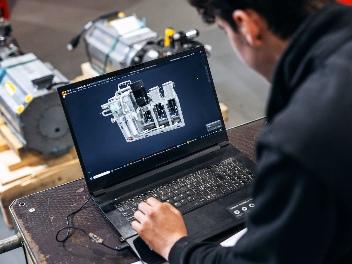 A person in a black sweater works on a laptop showing a 3D mechanical model on screen, with industrial machinery in the background.