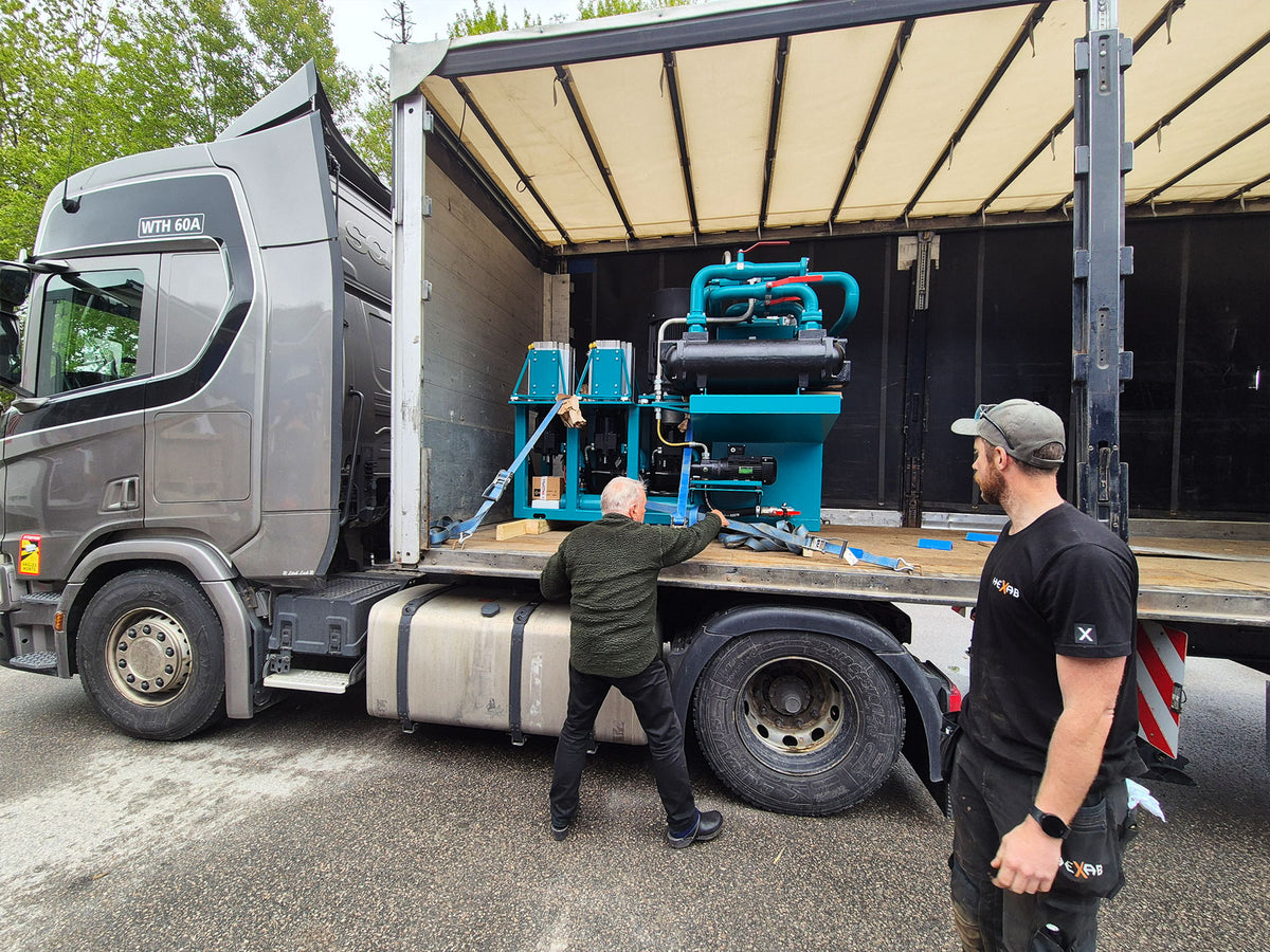 A blue industrial hydraulic machine getting loaded onto a large gray semi-truck with its side open. Two men are present near the truck and machine in an outdoor lot.