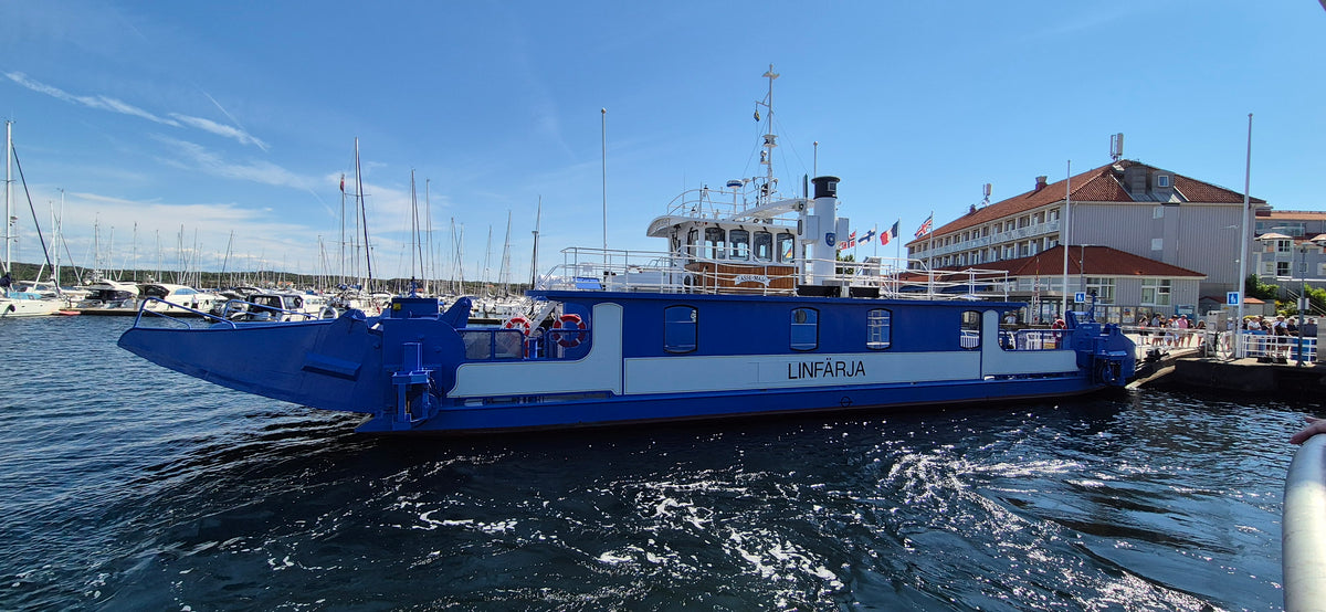 A blue and white ferry with the swedish word LINFÄRJA is shown docking to land with water, other boats, and a blue sky in the background.