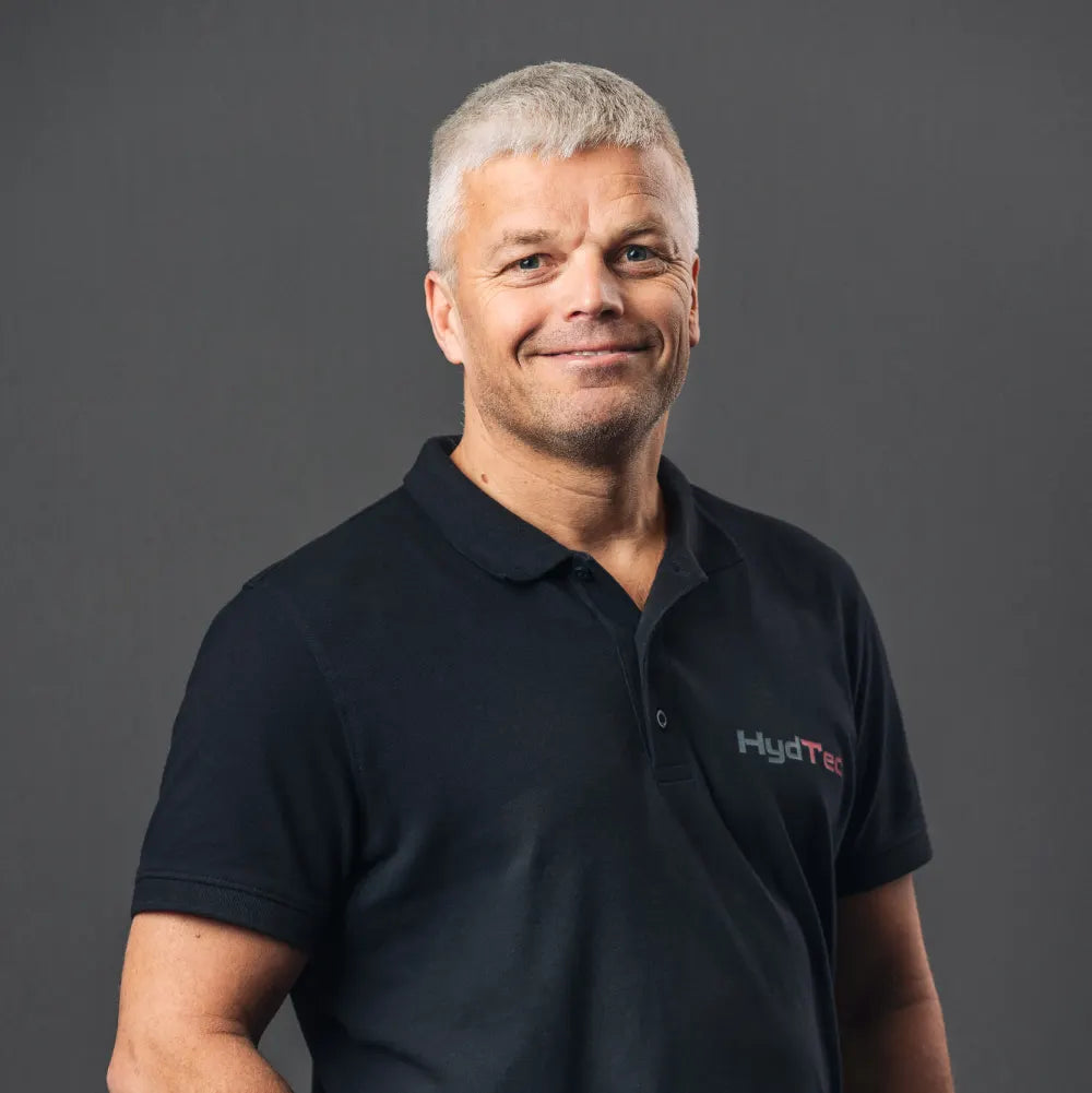 A man wearing a black short-sleeve polo shirt with a 'HydTec' logo on the left chest, set against a solid dark gray studio background.