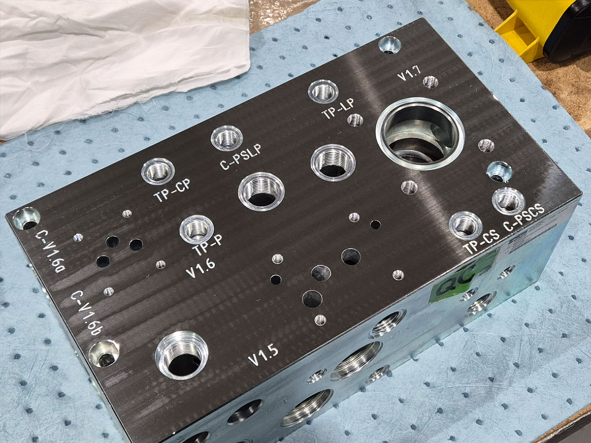 A dark metal hydraulic manifold block with numerous threaded ports and a large central cartridge valve bore on top. Ports are labeled with codes such as TP-CP, TP-LP, TP-P, TP-CS, C-PSLP, C-PSCS, and V1.5–V1.7. The block rests on a blue ESD-safe mat in a workshop setting.