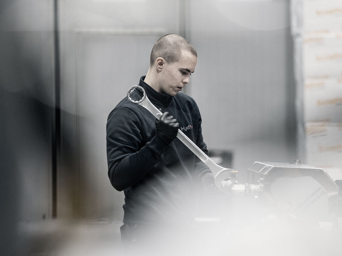HydTec employee in a black long-sleeve shirt and gloves holds a large silver wrench in an industrial setting with blurred machinery in the background.