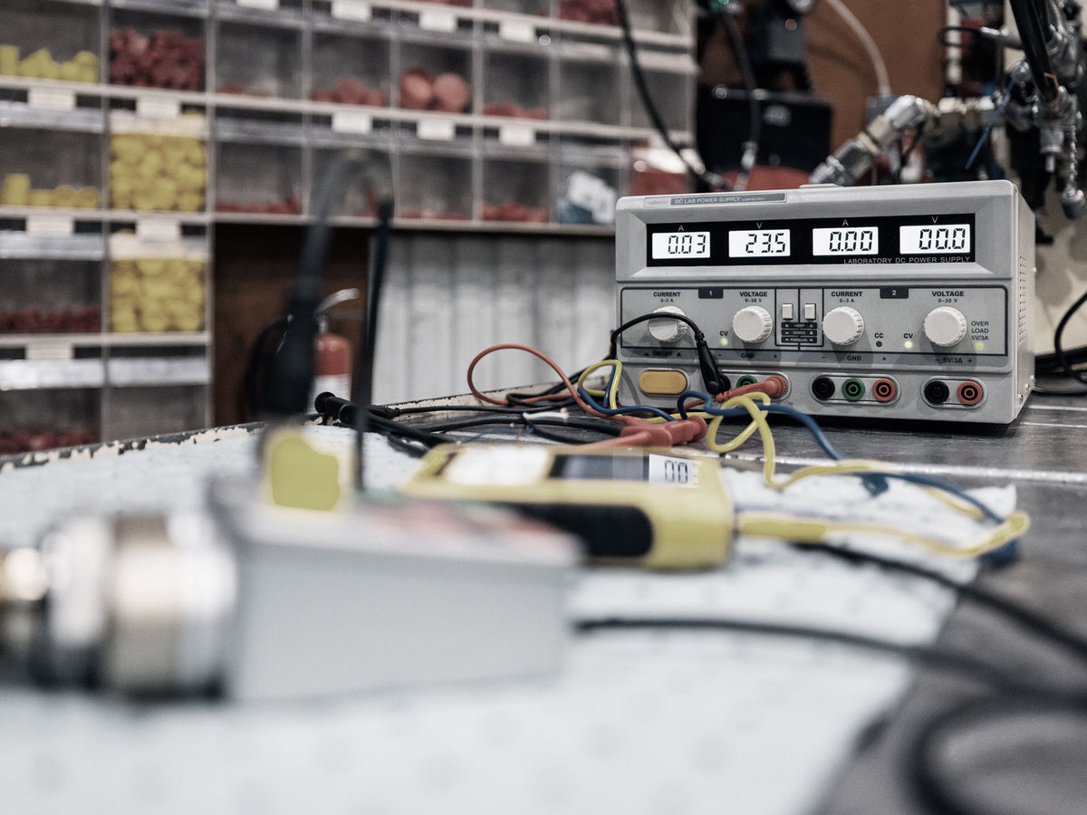A digital power supply with three display screens and rotary knobs sits on a workbench in a workshop, surrounded by wires, probes, and other electronic equipment. The background shows shelves filled with small parts.